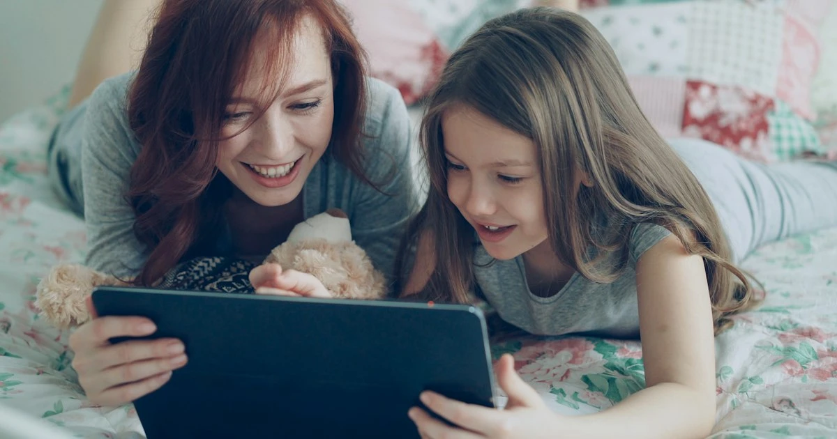 Mother and daughter lying on a bed, smiling at a tablet together